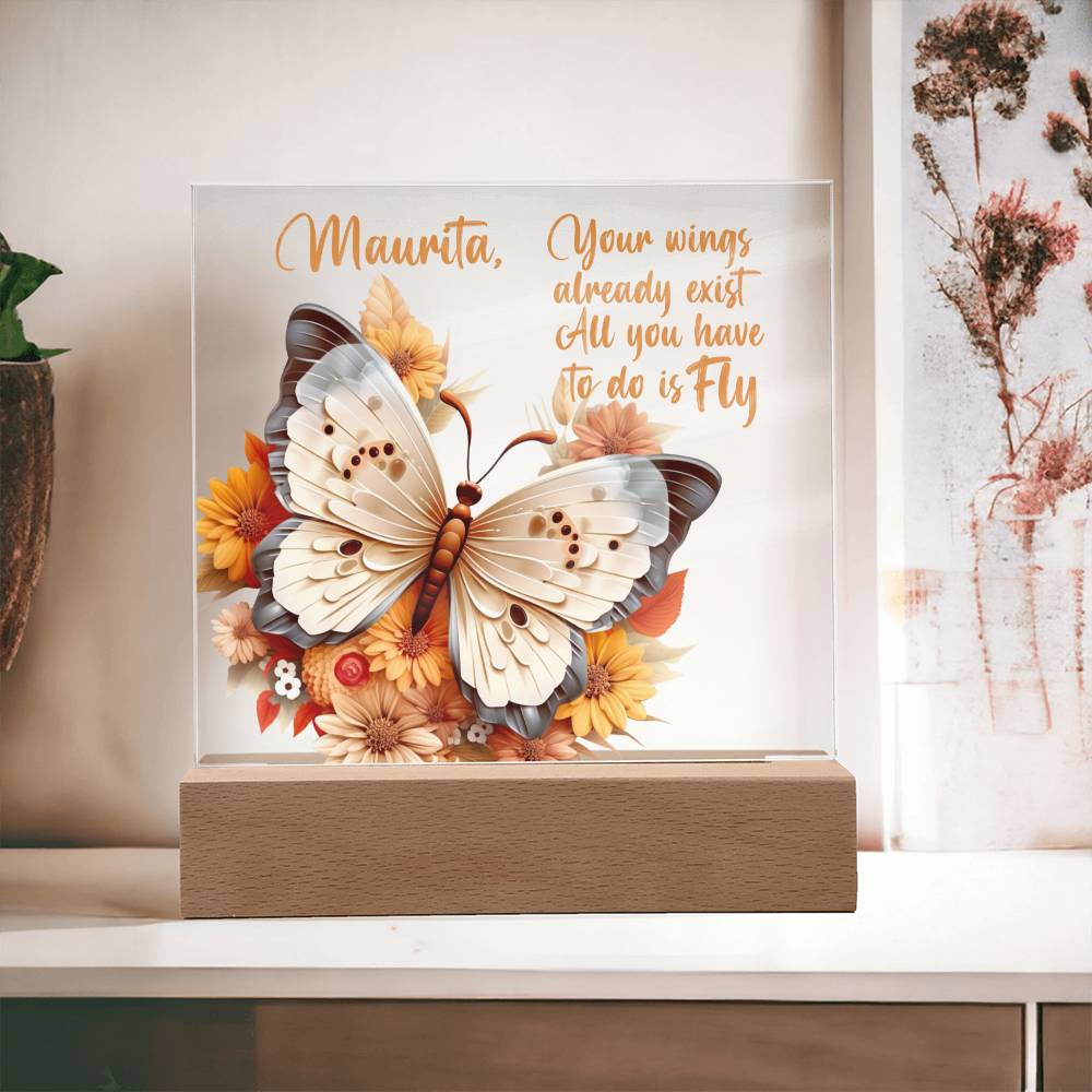 A clear acrylic plaque with a 3D butterfly in the colors of tan, beige and gray. Butterfly is surrounded by orange, yellow, peach, and tan flowers. Plaque sits in a light brown wooden base. Has a place to personalize name along with a message of encouragement. That reads "Your wings already exist All you have to do is fly."