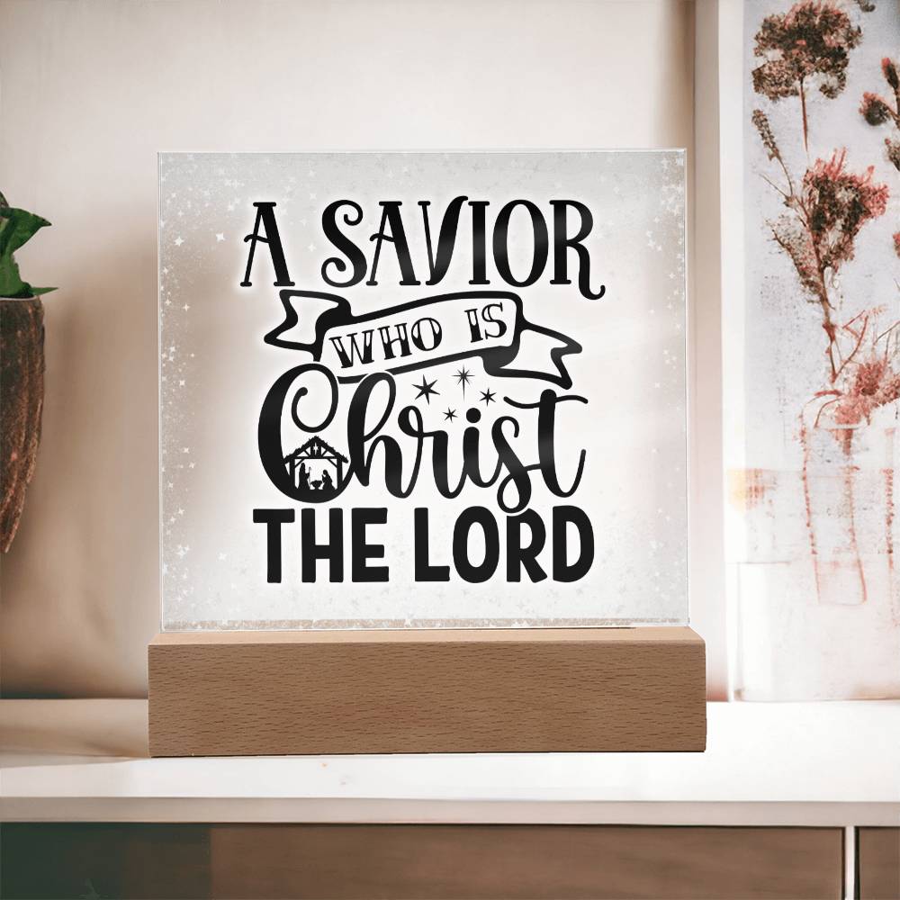 Christmas acrylic plaque with LED light wooden base. Plaque has the words A Savior Who Is Christ The Lord in a script font. An image of a nativity scene is nestled in the word Christ surrounded by stars.