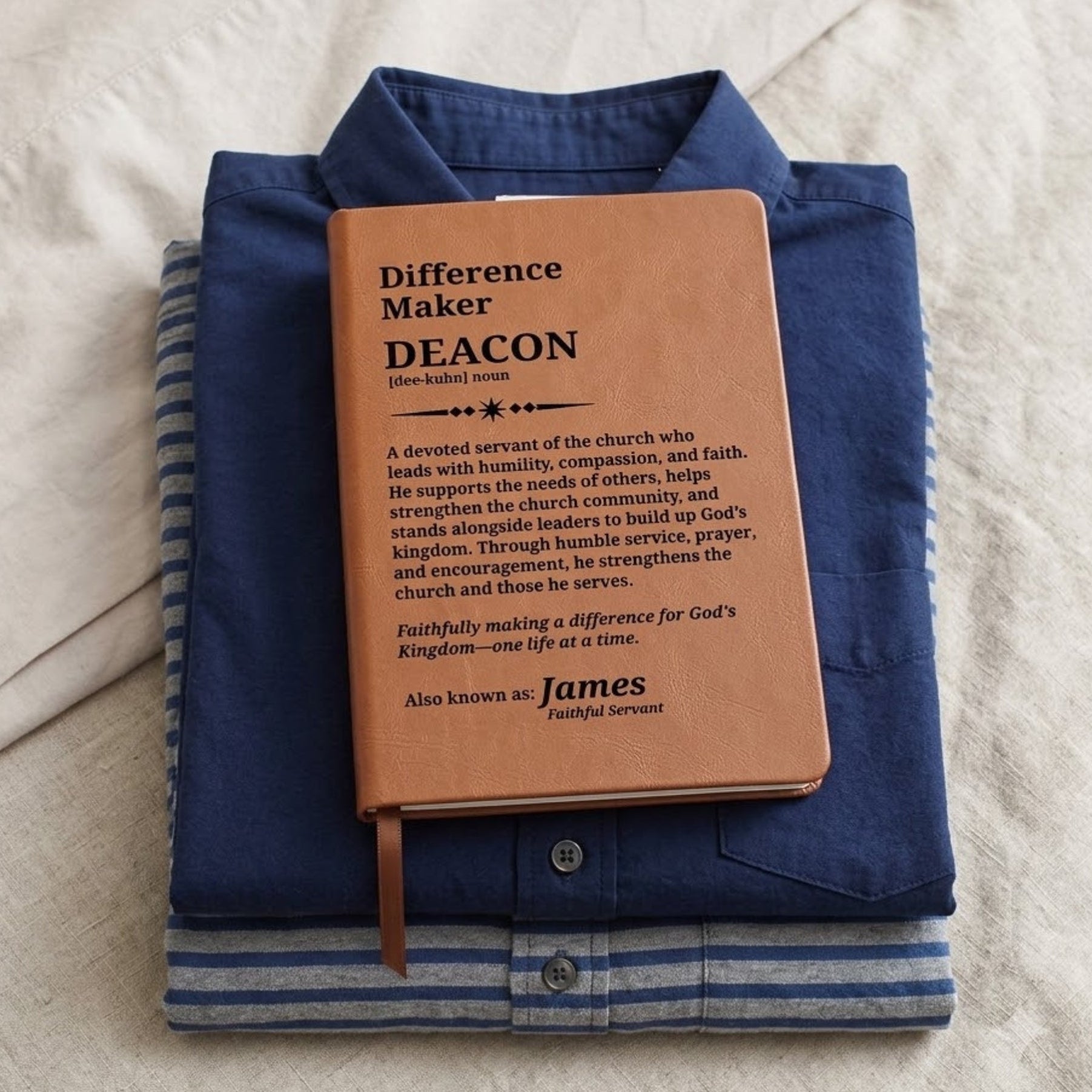 A personalized brown leather Bible journal with a bookmark ribbon, displayed on top of a folded blue striped button-down shirt on a textured white fabric background.