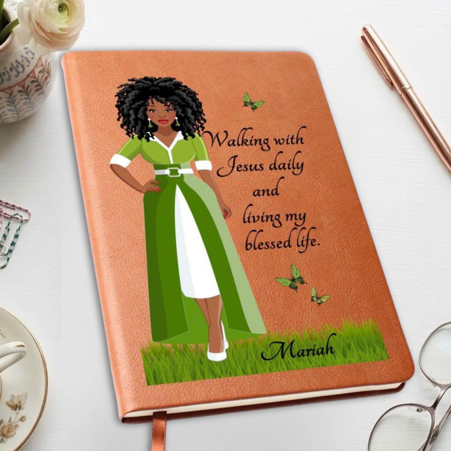 A personalized brown notebook with a textured cover, featuring an illustration of a Black woman in a green and white dress with the quote 'Walking with Jesus daily and living my blessed life.' and the name 'Mariah' at the bottom.