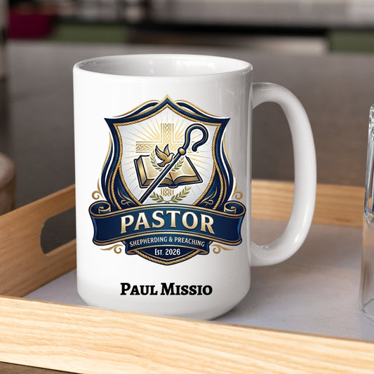 A white ceramic coffee mug with a custom pastor-themed crest graphic in navy blue, gold, and white, featuring the text 'PASTOR', 'SHEHERDING & PREACHING', and personalized name, 'PAUL MISSIO'. The mug is sitting on a light wood tray.