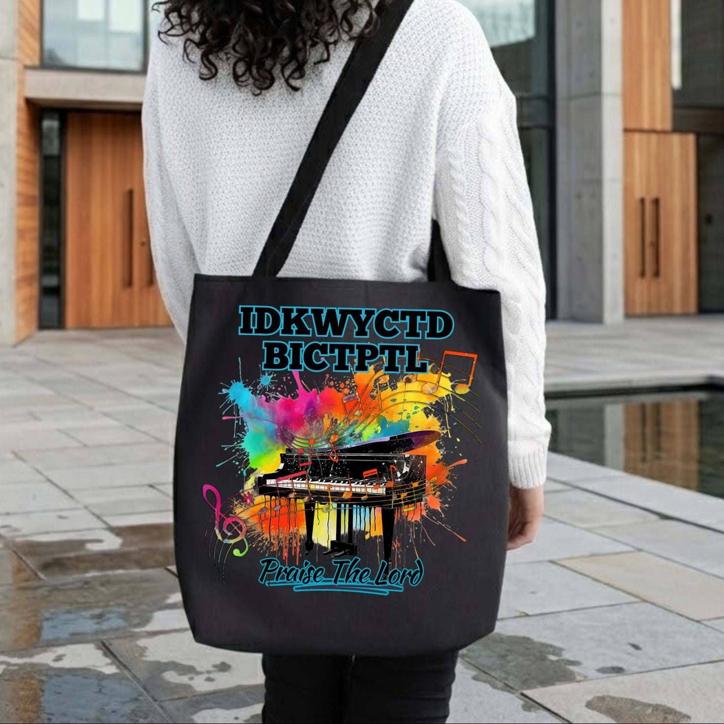 Lady standing in front of a modern church building holding a black tote bag with a colorful piano design and the text IDKWYCTDBICTPTL - Praise The Lord. 