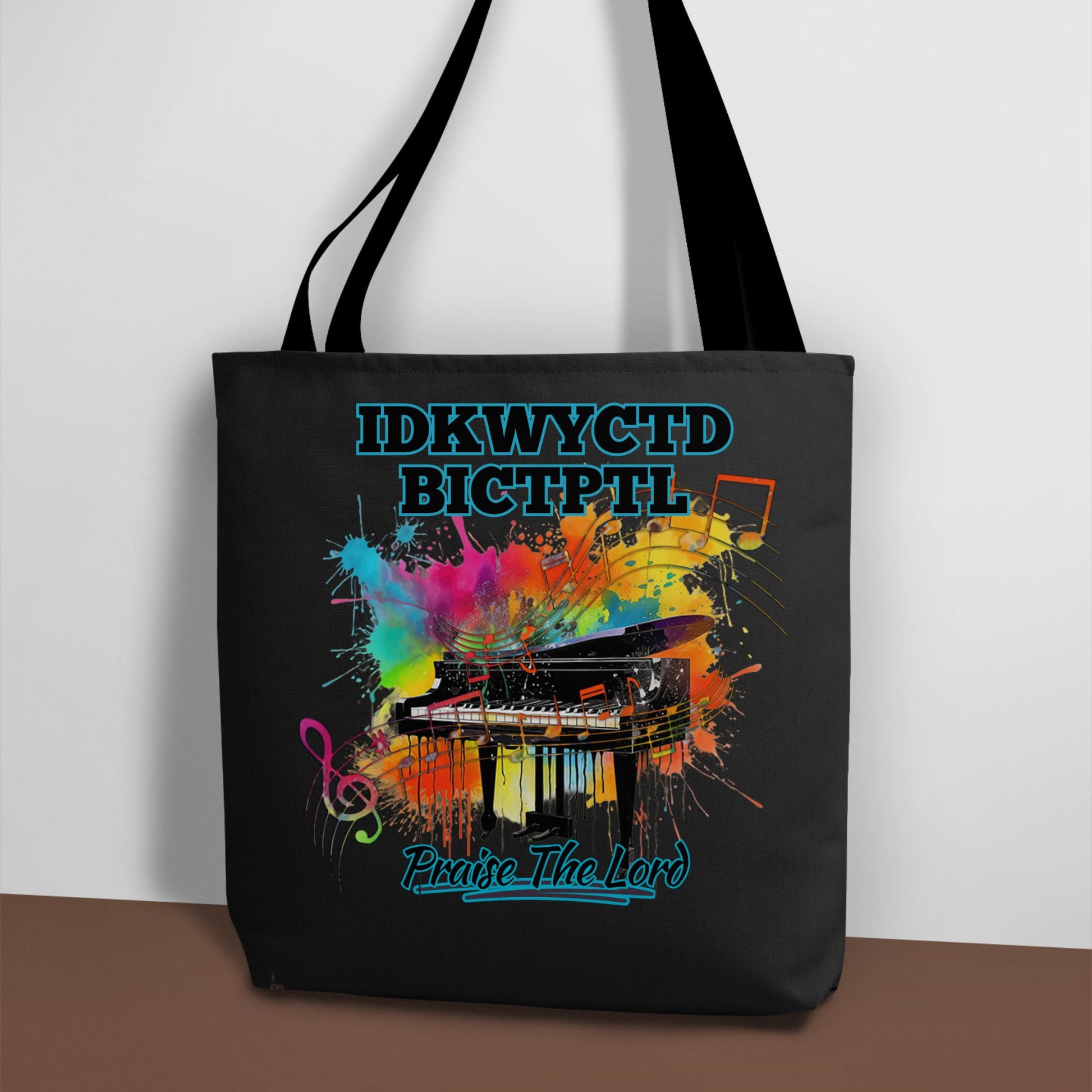 A black tote bag with a bright and colorful piano design and the text KWYCTDBICTPTL - Praise the Lord.