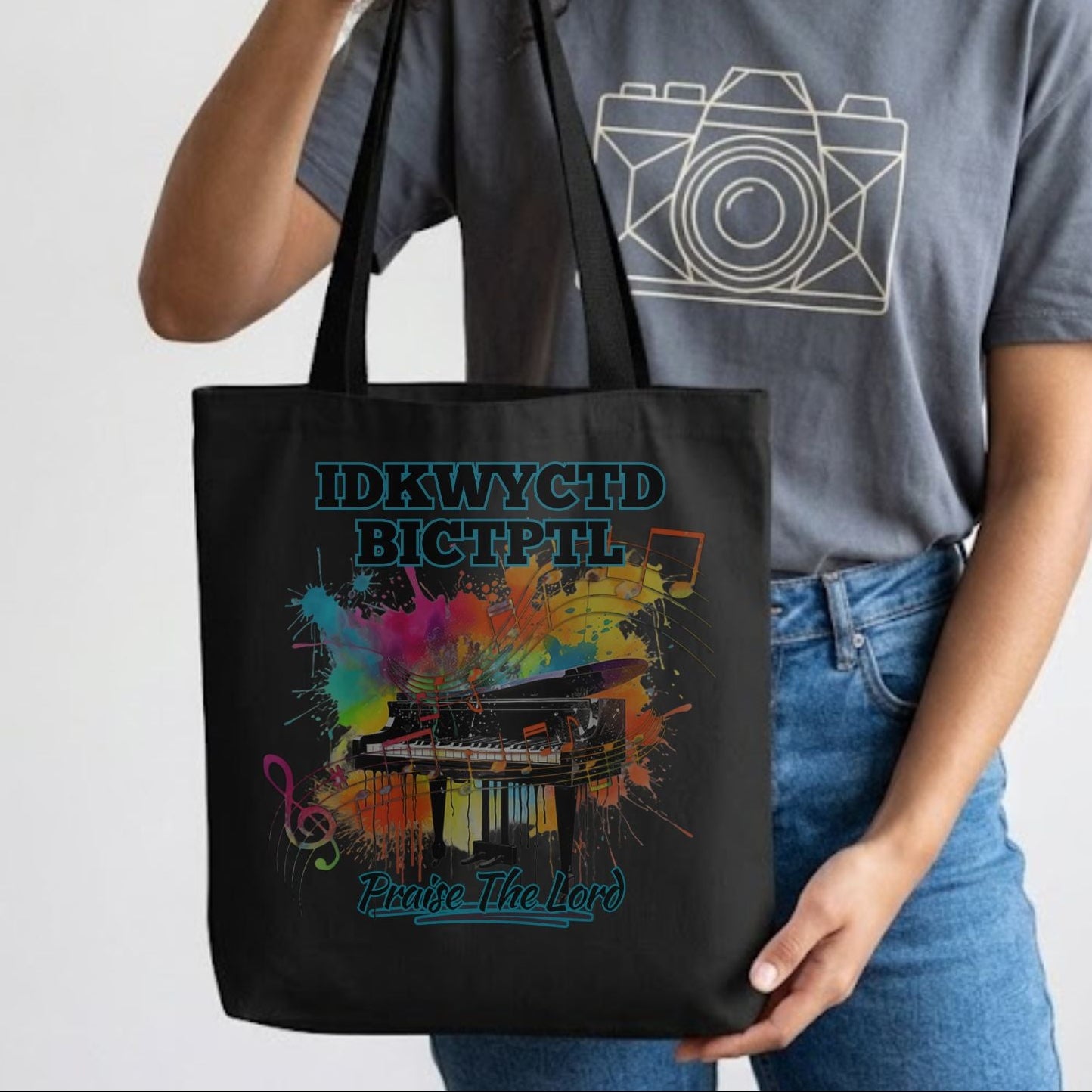 Lady holding a black tote bag with a colorful piano design and the text IDKWYCTDBICTPTL - Praise The Lord. 