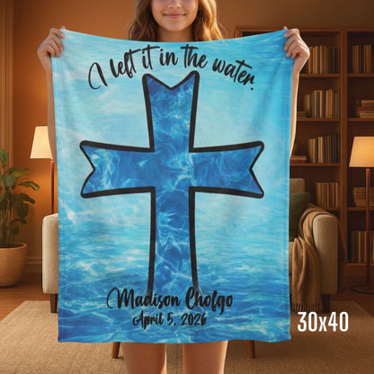 A person standing in a living room holding a small blue Sherpa baptism blanket that has an image of a cross in the water.
