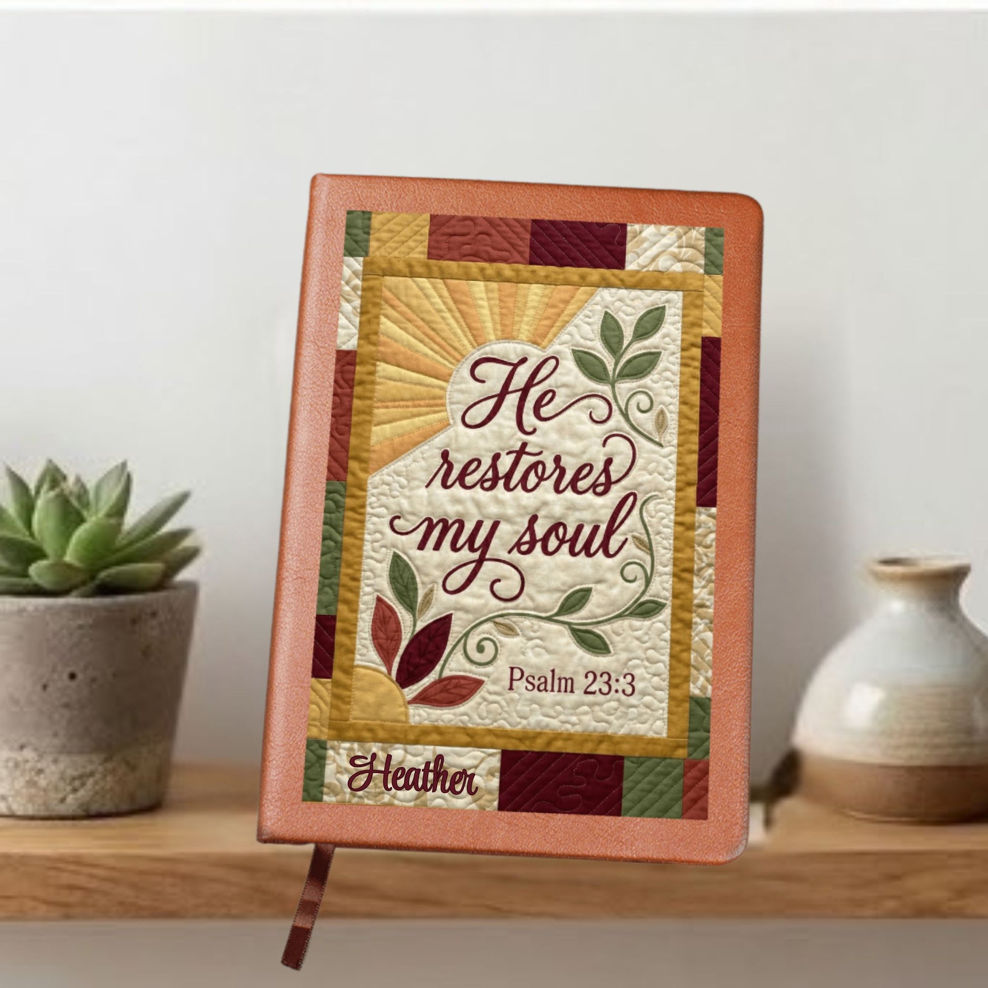 Vegan leather, faux quilt writing journal with text that reads- He Restores My Soul Psalm 23:3. Front view with plant.