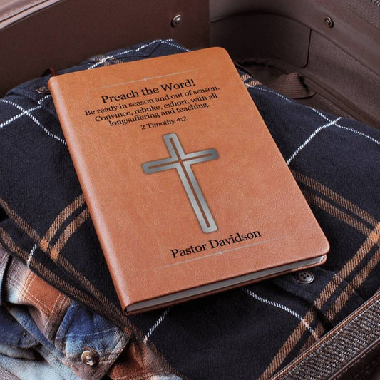 Brown vegan leather journal with a silver cross on the front cover packed in a suitcase. Front cover also features Bible verse 2 Timothy 4:2 as well as a place to personalize a name underneath the cross. Bible scripture and name is printed in black.