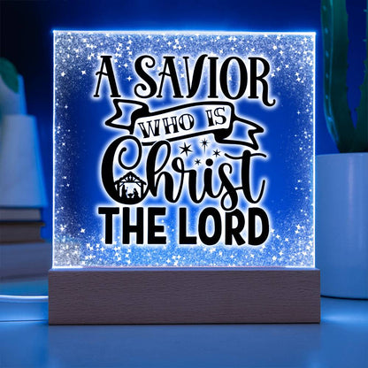 Christmas acrylic plaque with LED light wooden base. Plaque has the words A Savior Who Is Christ The Lord in a script font. An image of a nativity scene is nestled in the word Christ surrounded by stars. This image is illuminated by a blue LED light.
