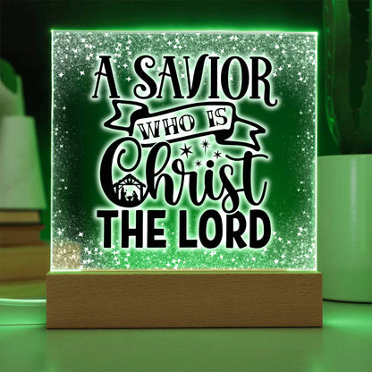 Christmas acrylic plaque with LED light wooden base. Plaque has the words A Savior Who Is Christ The Lord in a script font. An image of a nativity scene is nestled in the word Christ surrounded by stars. This image is illuminated by a green LED light.