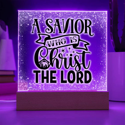 Christmas acrylic plaque with LED light wooden base. Plaque has the words A Savior Who Is Christ The Lord in a script font. An image of a nativity scene is nestled in the word Christ surrounded by stars. This image is illuminated by a purple LED light.
