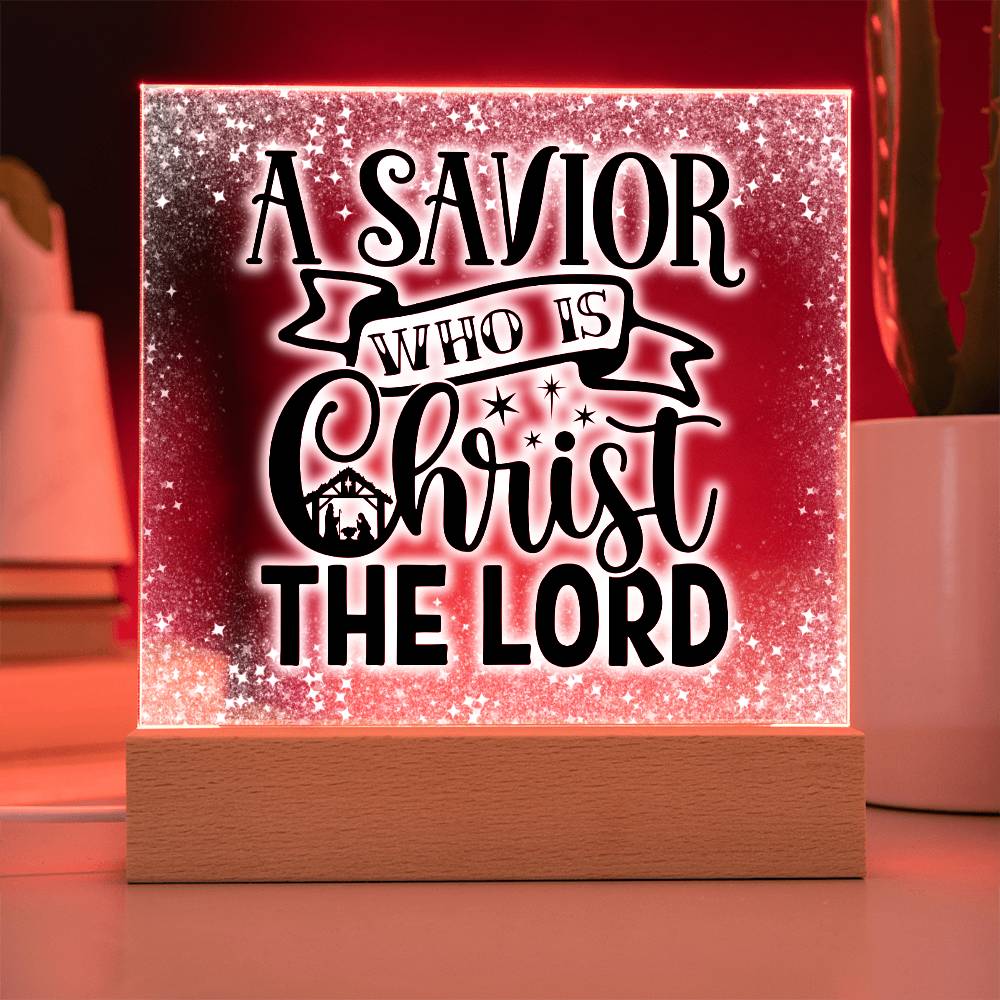 Christmas acrylic plaque with LED light wooden base. Plaque has the words A Savior Who Is Christ The Lord in a script font. An image of a nativity scene is nestled in the word Christ surrounded by stars. This image is illuminated by a red LED light.