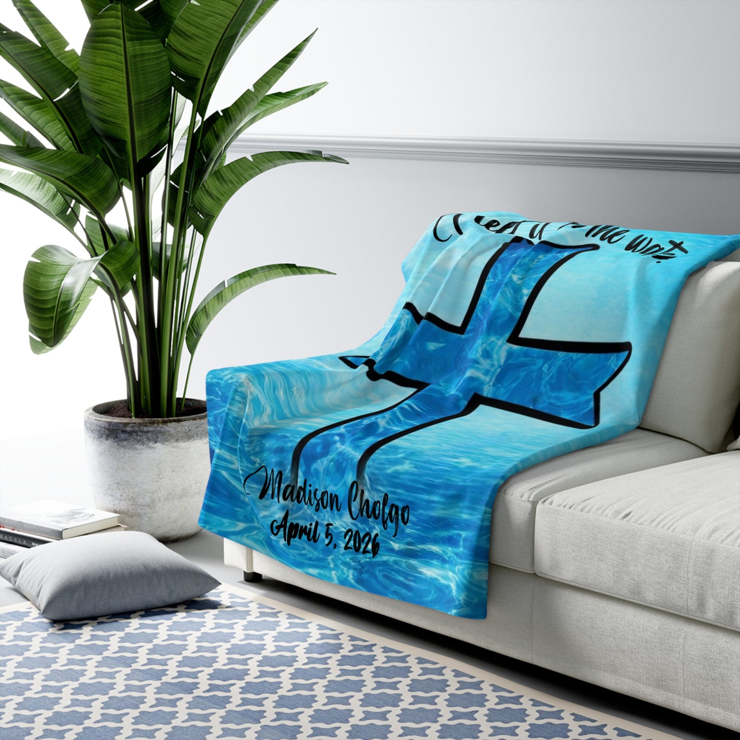 A blue Sherpa baptism blanket that has an image of a cross in the water, draped over a white couch, next to a potted plant.