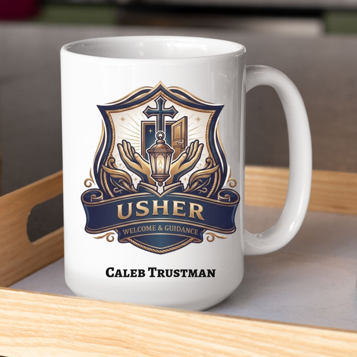 A white ceramic coffee mug with a custom printed crest design in navy blue, gold, and beige, featuring the text 'USHER' and 'WELCOME & GUIDANCE' with the personalized name 'CALEB TRUSTMAN' printed below. The mug is sitting on a light wood tray next to a clear glass tumbler.
