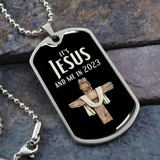 It's Jesus and Me In 2023 Christian dog tag necklace. Necklace is black with cross made of wood and a crown of thorns on top of it. Necklace is resting on flat rocks.