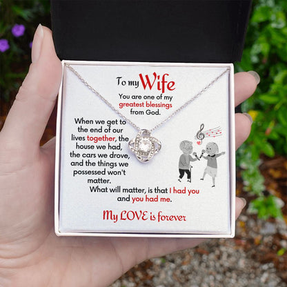 To Wife Love Knot necklace with jewelry message card. Card features special message to wife with an elderly man and woman singing together. Card is white background. Necklace in box being handheld.