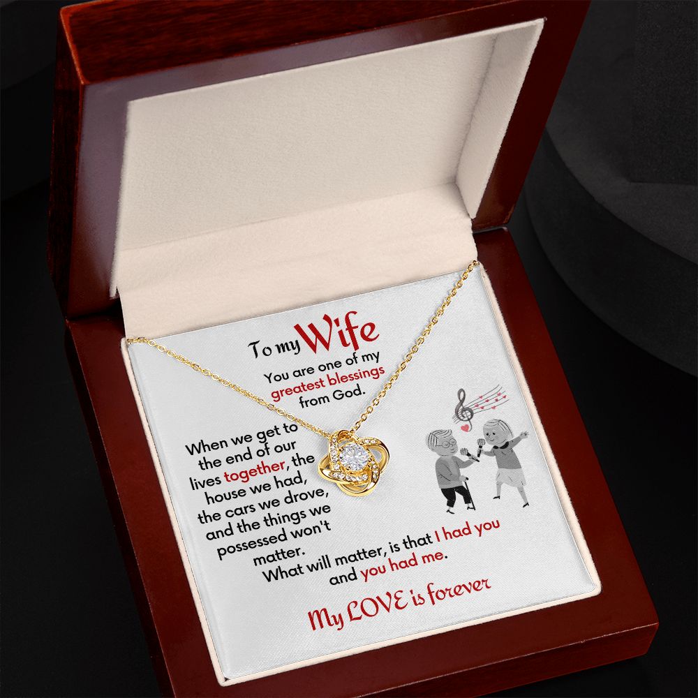 To Wife 18k yellow gold finish Love Knot necklace with jewelry message card. Card features special message to wife with an elderly man and woman singing together. Card is white background. In a mahogany box open.