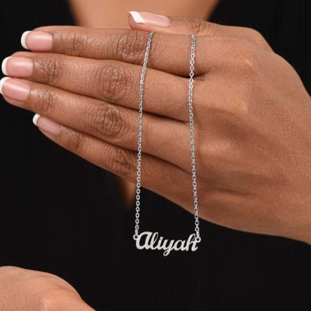 American Stainless Steel custom name necklace. The scripted name necklace being held in a hand in this image is "Aliyah." Supports up to 8 characters for the name necklace. Name height measures 0.3".